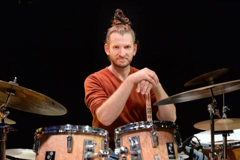 Mann mit Dutt und rotem Oberteil spielt Schlagzeug, hält Drumsticks, im Hintergrund weitere Trommeln und Becken, dunkler Hintergrund.