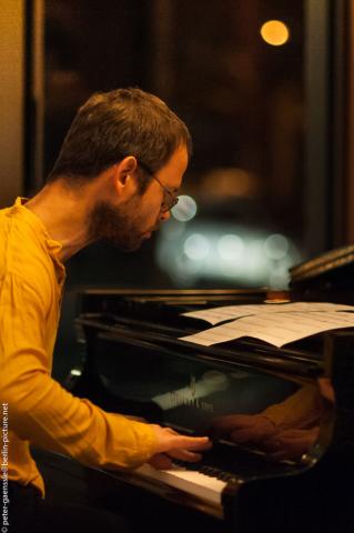 Mann mit Brille und gelbem Oberteil spielt Klavier, Notenblätter auf dem Flügel, unscharfe Lichter im Hintergrund, konzentrierte Haltung.