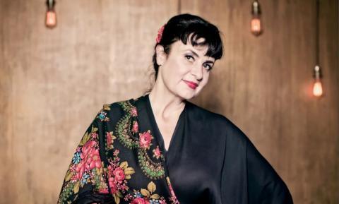 Frau mit kurzem schwarzem Haar und roter Blume im Haar, trägt schwarzes Kleid mit buntem Blumenmuster, lächelnd, Holzvertäfelung und zwei Wandlampen im Hintergrund.
