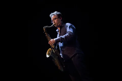 Mann mit Brille und blauem Hemd spielt goldfarbenes Saxophon, konzentriert, dunkler Hintergrund, seitliche Beleuchtung betont Instrument und Hände.