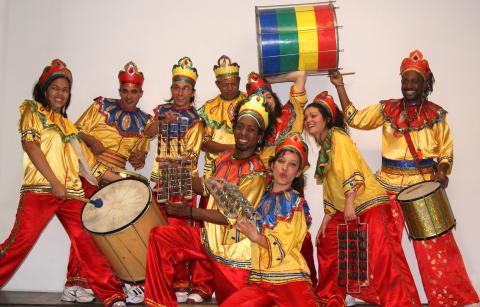 Acht Musiker in gold-rot-blauen Kostümen mit buntem Kopfschmuck, posierend mit Trommeln, Rasseln und einer grün-gelb-roten Trommel.