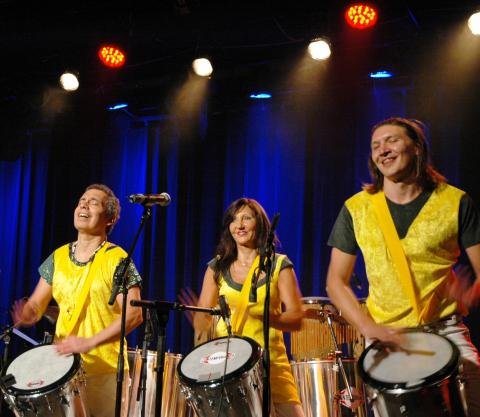 Drei Musiker in gelben Westen spielen Trommeln auf der Bühne, Mikrofone vor ihnen, blaue Vorhang-Hintergrundbeleuchtung mit Spotlights.