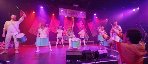 Sieben Musiker in weißen Outfits spielen Trommeln und Becken auf einer pink beleuchteten Bühne mit transparenten Vorhängen.