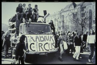 Schwarz-weiß Foto mit LKW der 60er Jahre, auf der Front ein bemaltes Transparent mit der Aufschrift "Fabrik Circus". Drum herum und auf dem Wagen stehen und laufen junge Leute mit langen Haaren.