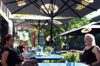 Outdoor seating area of the Rudi & Rosa restaurant at the ufaFabrik, featuring people enjoying a sunny day under large black umbrellas with the Wurstbraterei logo. Blue tables are adorned with small potted plants, and greenery surrounds the relaxed setting.