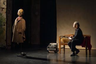 Bühnenbild: Links eine Puppe in braunem Mantel und kariertem Hemd, rechts eine sitzende Person in Schwarz am Tisch mit Akkordeon und Fächer, dunkler Hintergrund mit Lichtakzenten.
